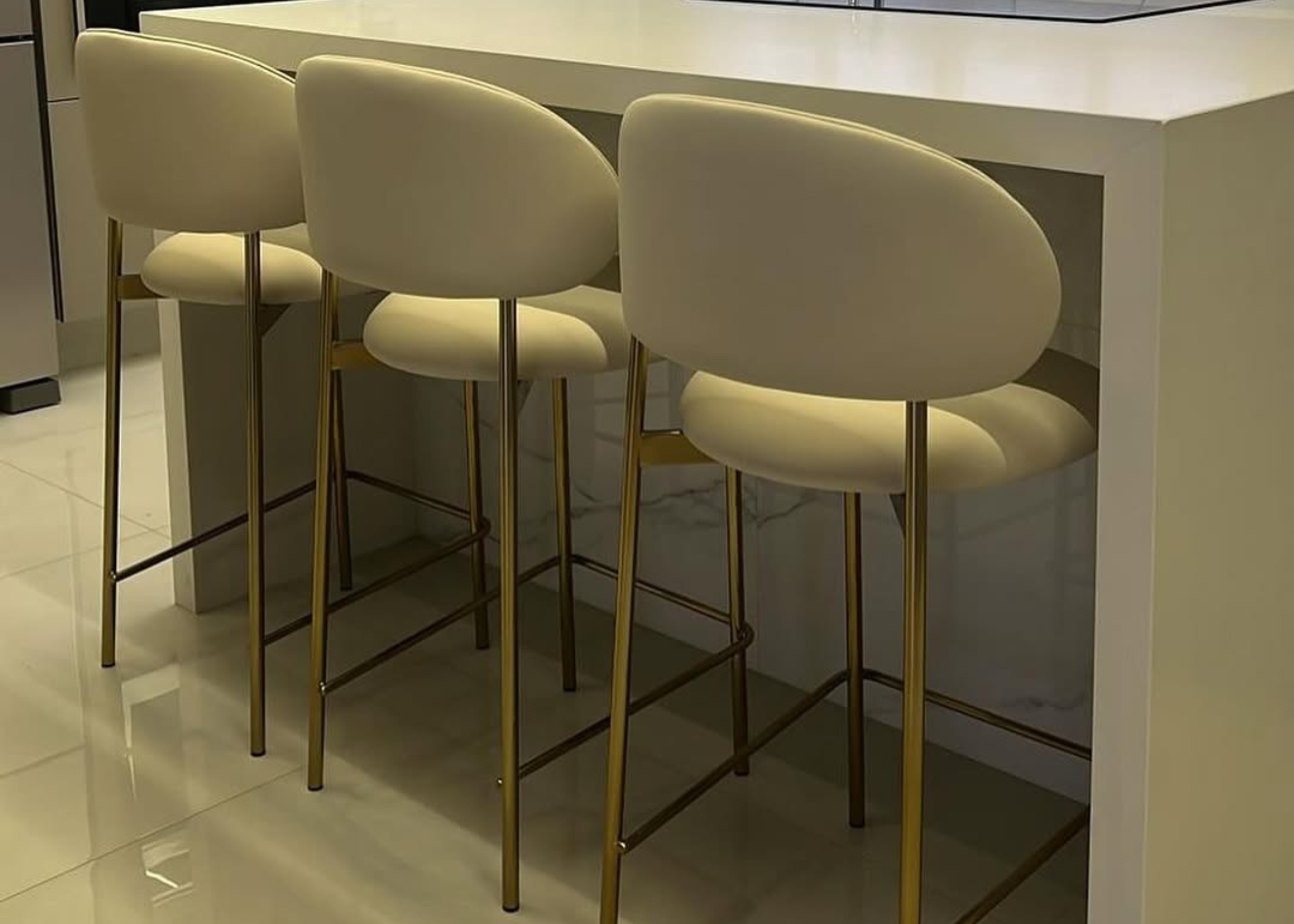 Banqueta Doralta com base dourada e estofado em tecido off-white, ambientada em cenário moderno. Móvel sofisticado e funcional para bancadas e áreas gourmet elegantes.