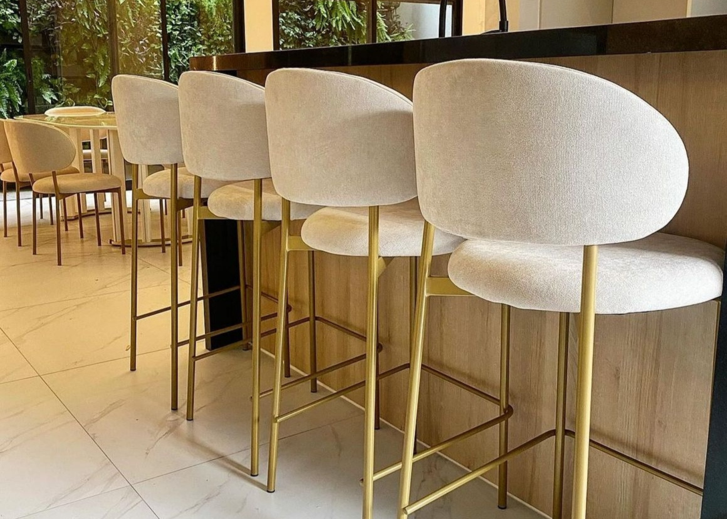 Banqueta Doralt estofada em tecido off-white com base dourada, disposta em bancada moderna. Móvel elegante e funcional para áreas gourmet sofisticadas.