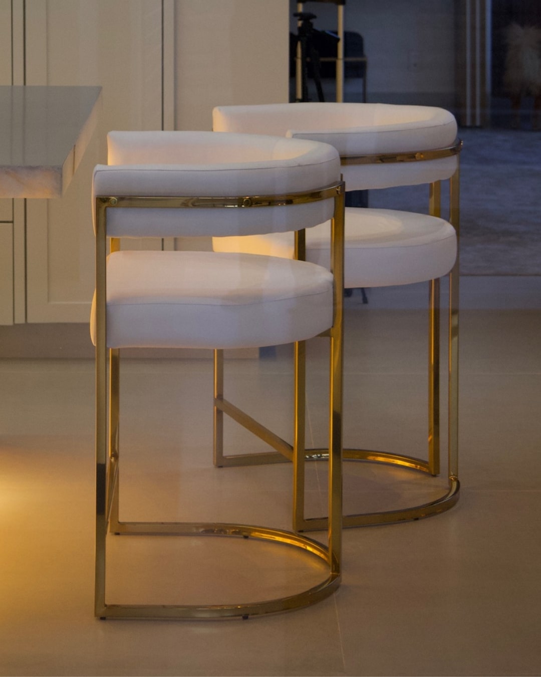 Banquetas de luxo Eldorado com estofado branco e estrutura dourada em ambiente de alto padrão.