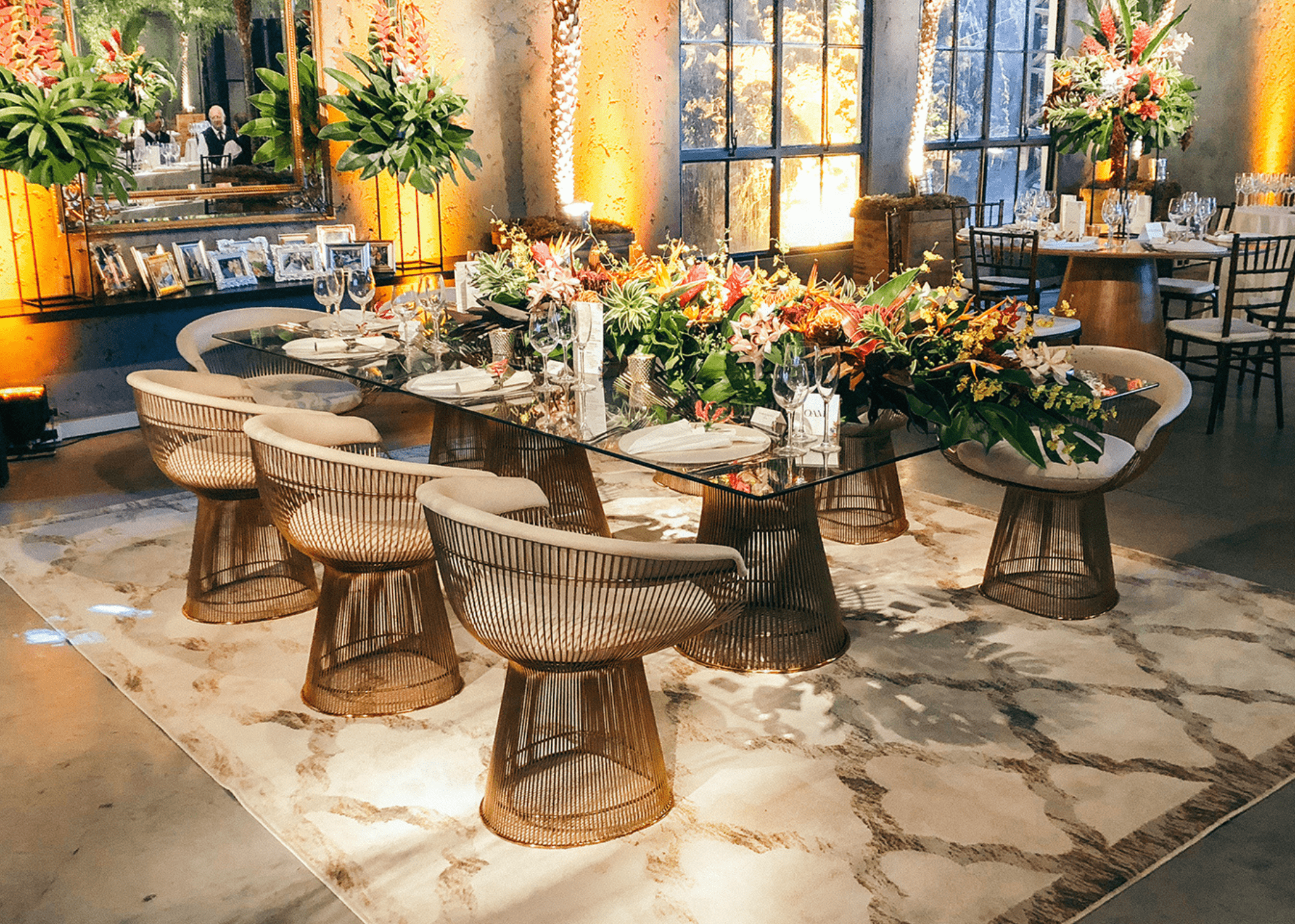 Cadeira Platner em aço inox e estofado branco em sala de jantar sofisticada, com mesa de vidro e decoração floral luxuosa.