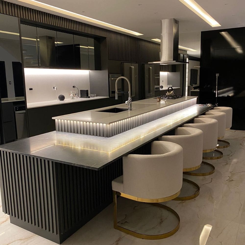 Cozinha gourmet contemporânea com ilha central e bancada iluminada, acompanhada por poltronas Gregori com encosto curvo estofado e base em aço inox dourado. Composição sofisticada e funcional com mobiliário de alto padrão da Moveca Decor.