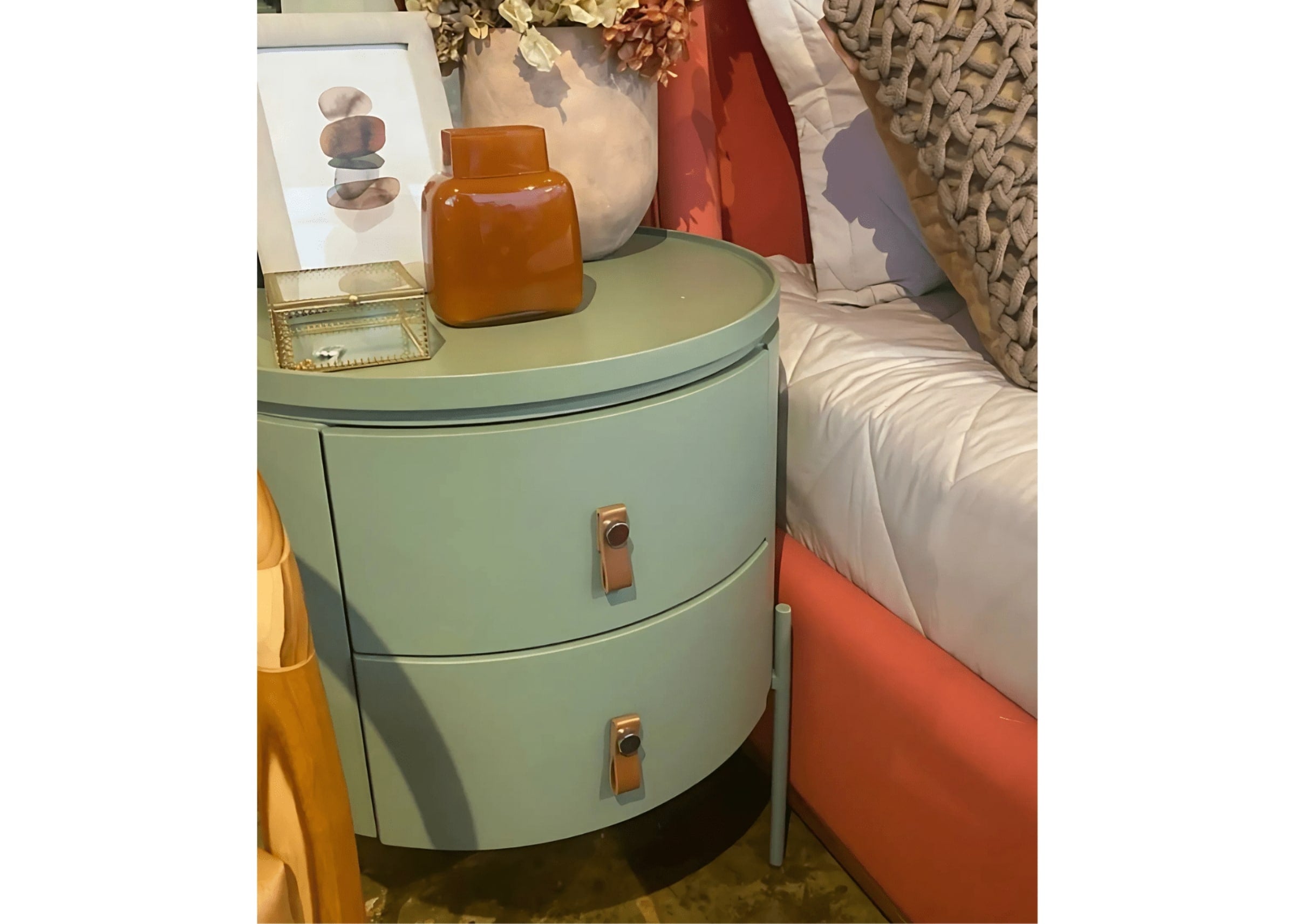 Mesa de cabeceira redonda verde menta com dois gavetões e puxadores em couro ao lado da cama.