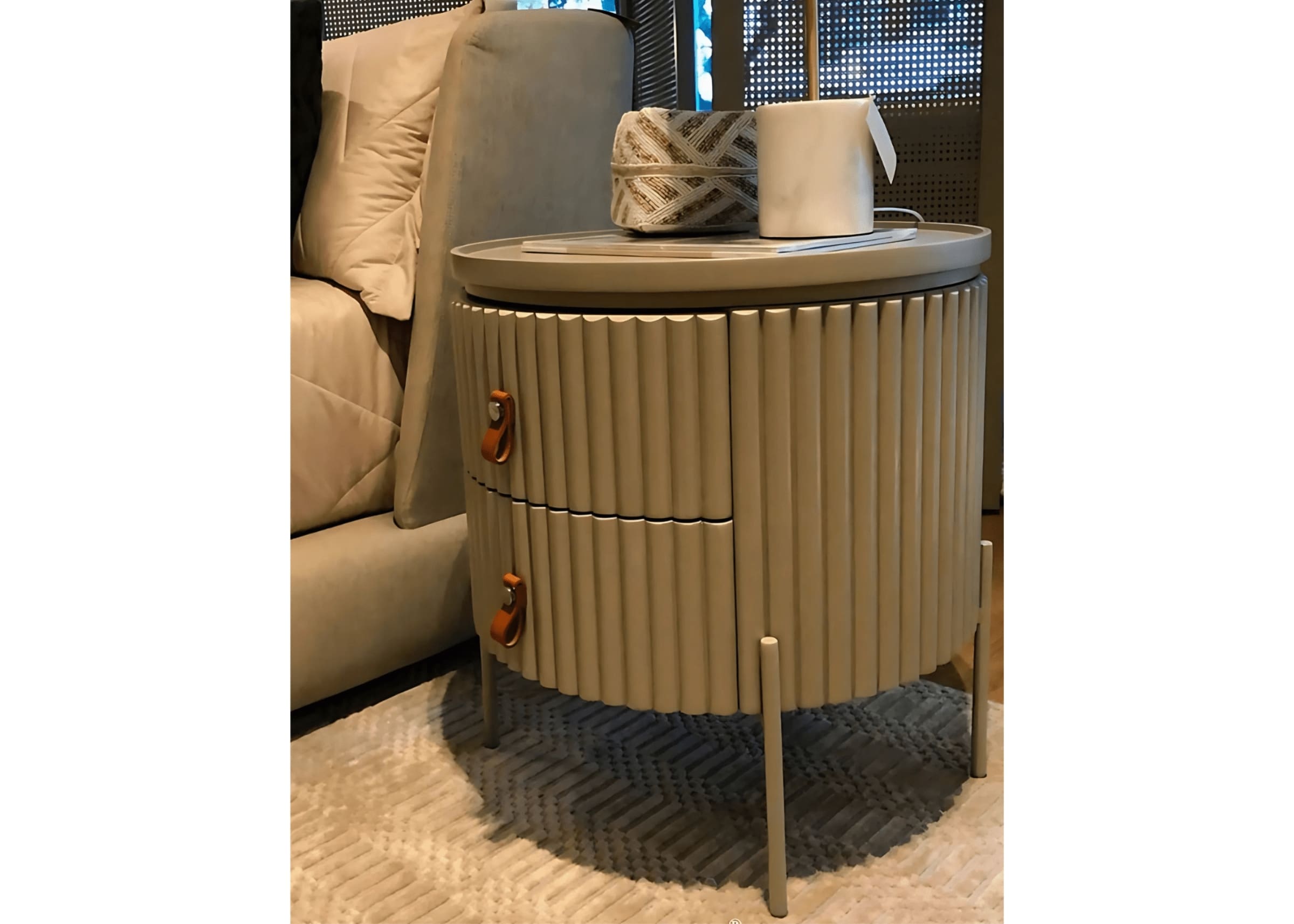 Mesa de cabeceira redonda com acabamento ripado fendi e alça de couro caramelo, ao lado da cama.