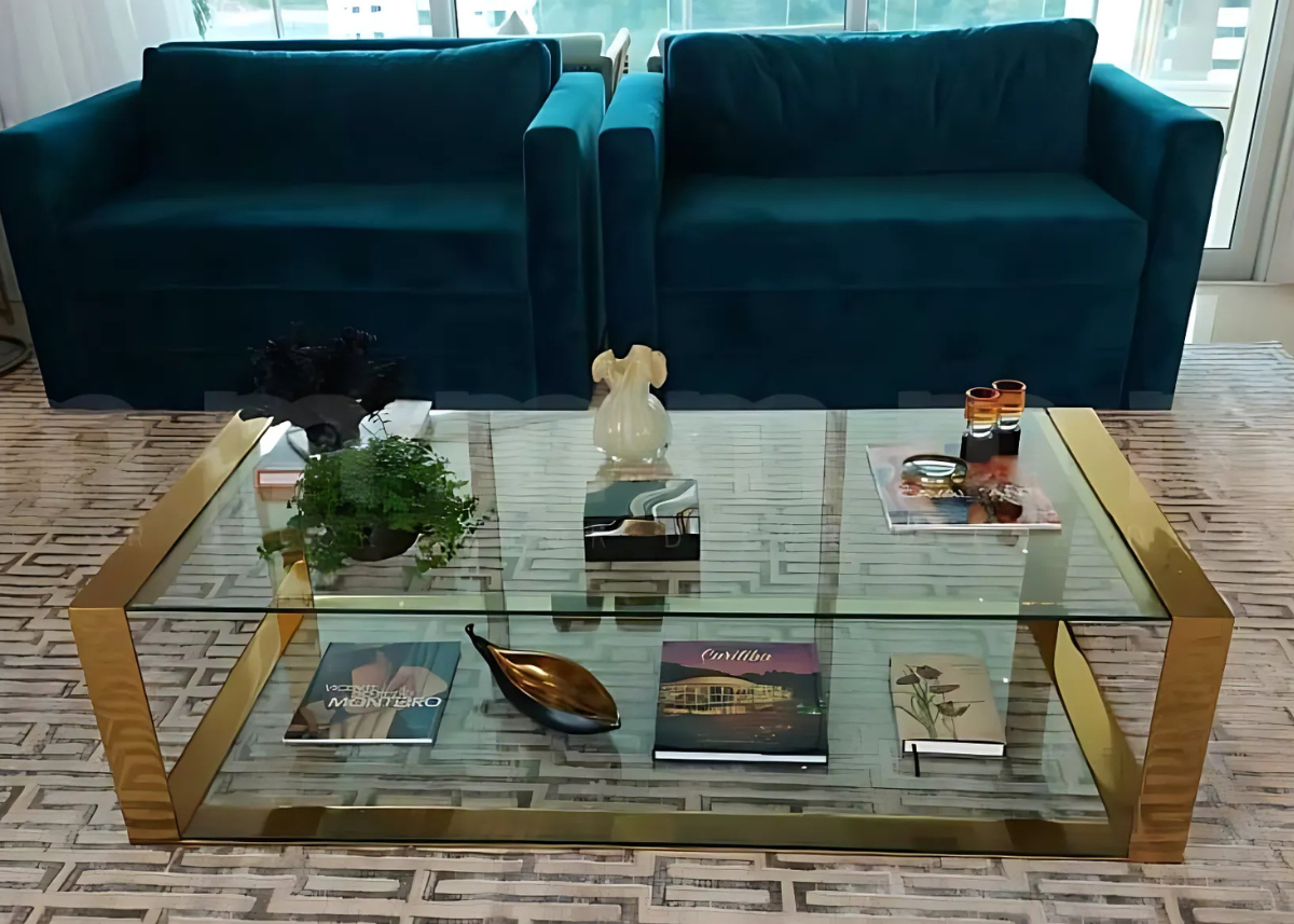 Mesa de centro com tampo de vidro transparente e base em inox dourado, vista em sala de estar moderna. Peça elegante para compor ambientes sofisticados e iluminado.