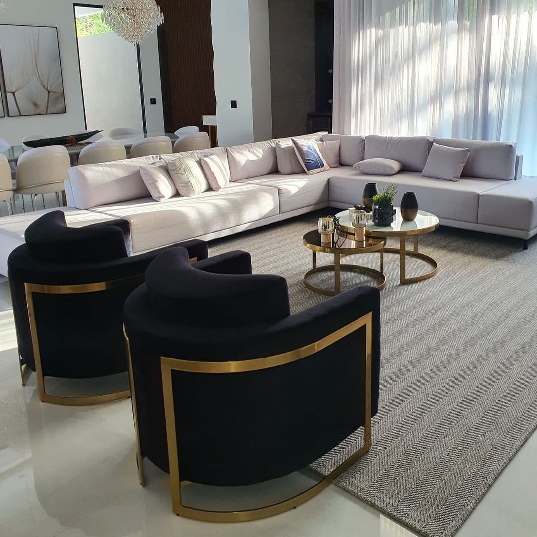 Sala de estar luxuosa com poltronas Eldorado em veludo preto com estrutura em aço inox dourado, mesa de centro Vegas com base metálica gold e tampo espelhado, e sofá em L off-white com puff. Composição elegante com móveis de alto padrão da Moveca Decor.