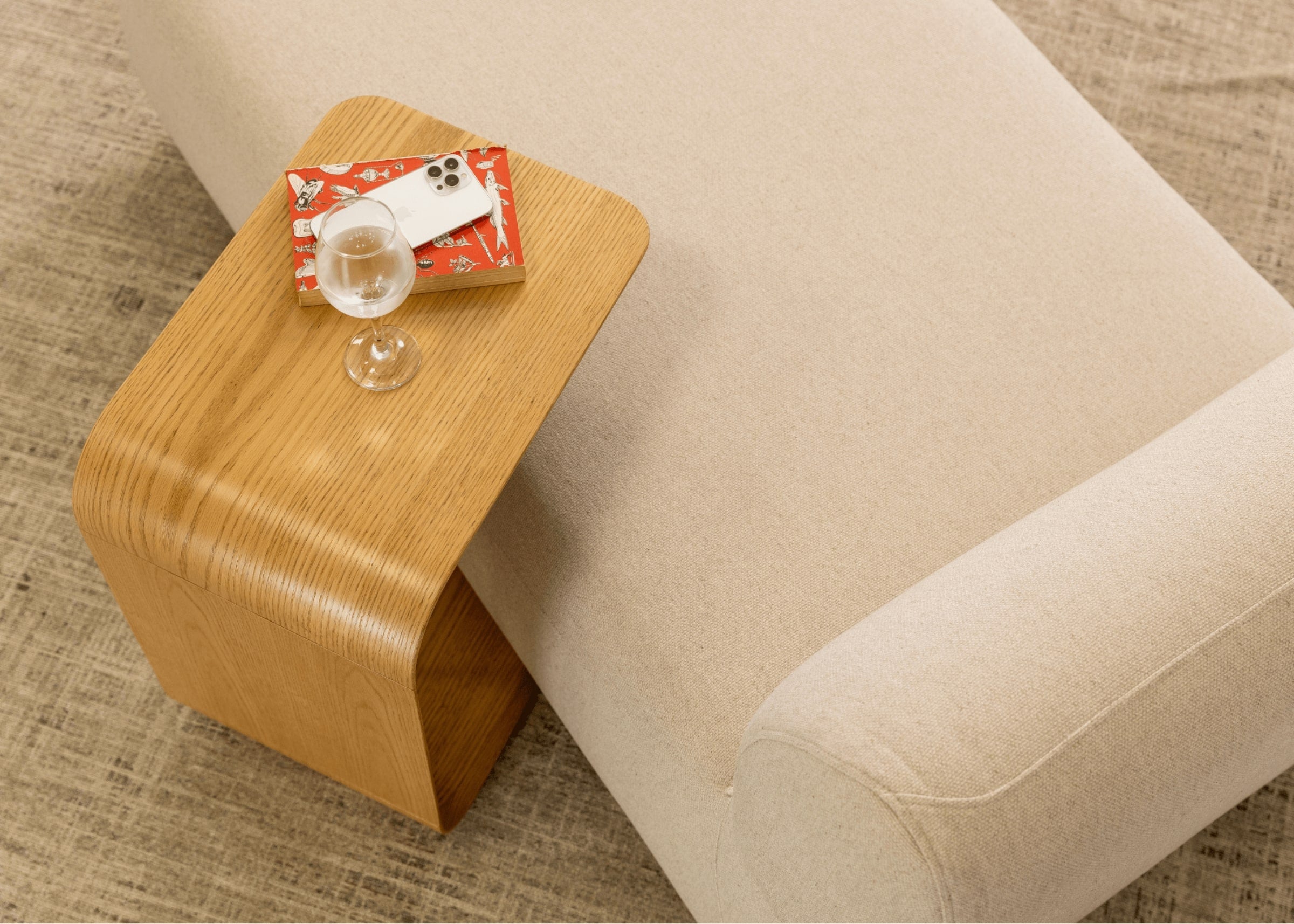 Mesa lateral em formato C ao lado de sofá, usada como apoio para livro, taça e celular. - Mesinha lateral Madri